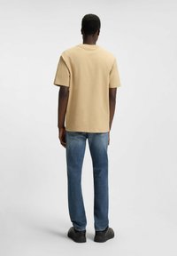 T-shirt in cotone beige, vestibilità casual, scollo rotondo, maniche corte, abbinato a jeans in denim blu, con taglio dritto e dettagli minimi.