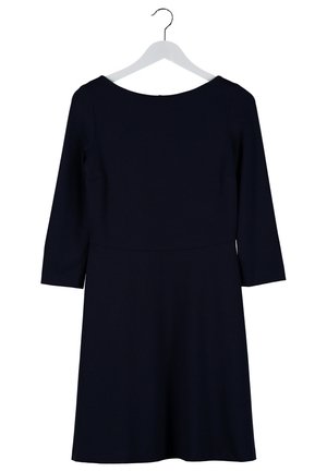Mujer con un vestido negro hasta la rodilla con mangas tres cuartos, sosteniendo gafas de sol con montura carey en una mano, y llevando sandalias negras con detalles dorados.