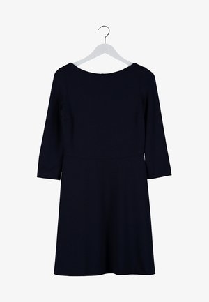 Mujer con un vestido negro hasta la rodilla con mangas tres cuartos, sosteniendo gafas de sol con montura carey en una mano, y llevando sandalias negras con detalles dorados.