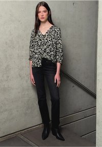 Blusa floreale in bianco e nero con scollo a V e maniche a tre quarti, abbinata a jeans scuri e stivali neri alla caviglia, posata su uno sfondo di cemento.