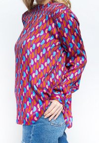 Blusa colorata con colletto alto, maniche lunghe e un motivo a goccia in toni di rosa, blu e arancione, realizzata in tessuto morbido.