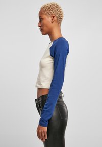 Kort T-shirt met een crème kleurige romp en blauwe lange mouwen, gemaakt van een katoenmengsel. Fitted ontwerp, zijaanzicht toont een gladde textuur en rechte snit.