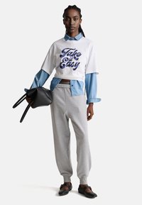 Bílé oversized tričko s potiskem "Take it Easy", vrstvená džínsová košile, šedé tepláky a černé baleríny, držící černou kabelku.
