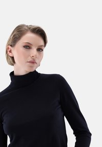 Schwarzer Rollkragenpullover in einer enganliegenden Design, mit einer glatten Textur. Das Model hat kurze blonde Haare und steht vor einem schlichten Hintergrund.