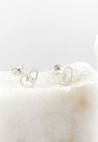 Silver hjärtformade örhängen med ett utdraget mönster inuti, med en slät yta och en polerad finish. Monterade på en plan yta.