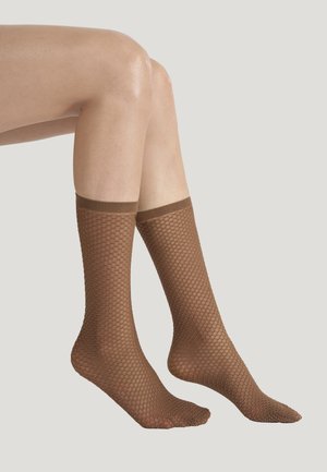 Benen met doorzichtige bruine maasachtige sokken met een diamantpatroon tegen een effen lichtgrijze achtergrond.