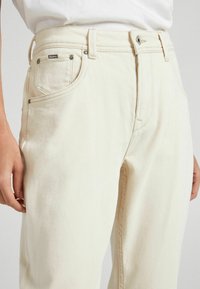 Lähikuva henkilöstä, joka pitää yllään korkeavyötäröisiä, kermaisen värisiä farkkuja, joissa on etutaskut ja Pepe Jeans -logo pienessä oikeassa taskussa.