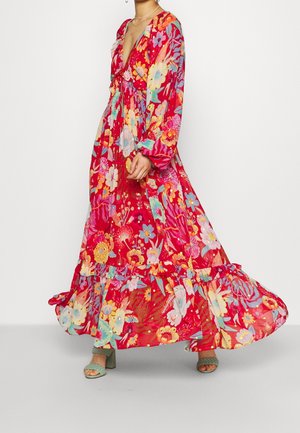 Femme portant une longue robe rouge fluide à fleurs avec manches longues et sandales vertes à talons sur un fond blanc uni.