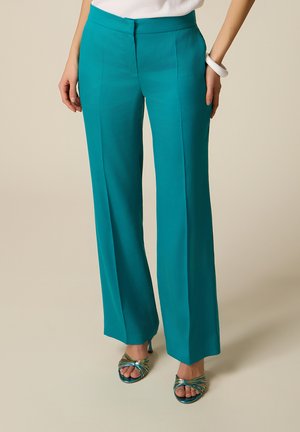 Donna che indossa pantaloni a gamba larga color teal con piega frontale, top bianco, braccialetto bianco e sandali aperti multicolore, in piedi su pavimento beige.