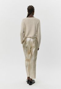 Bežinis megztinis su laisva forma, derinamas su blizgiais auksiniais tekstūruotais kelnais ir juodais batais, matomas iš nugaros.