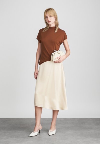 Top scurt cu mânecă brown, fustă midi din satin crem, încălțăminte albă cu detalii din dantelă, ținând o geantă de mână texturată crem.