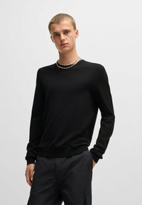 Maglione nero a maniche lunghe in una texture liscia, con colletto girocollo e polsini a costine, indossato con pantaloni cargo scuri e collane sovrapposte.