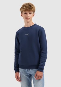 Tienerjongen in een marineblauwe sweatshirt met de tekst "BALLIN" en lichtblauwe jeans, staande tegen een effen lichte achtergrond.