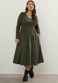 Kobieta w oliwkowej zielonej sukience midi z długim rękawem i zebranym w talii, stojąca z jedną ręką w kieszeni i czarnych sznurówkach.