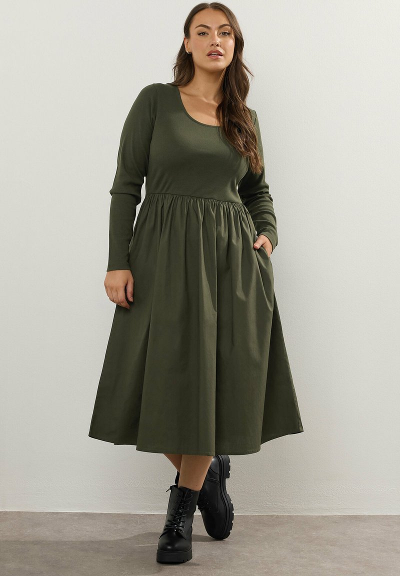 Kobieta w oliwkowej zielonej sukience midi z długim rękawem i zebranym w talii, stojąca z jedną ręką w kieszeni i czarnych sznurówkach.