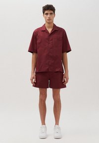 Camicetta corta marrone con una tasca sul petto e pantaloni abbinati. Il tessuto è liscio. Indossato con sneakers bianche, su uno sfondo chiaro.