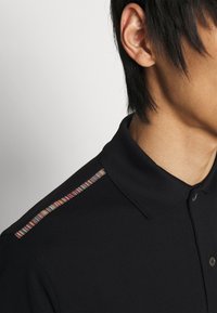 Svart polo som har ett texturerat tyg, korta ärmar och en färgglad randig detalj längs axelsömmen.