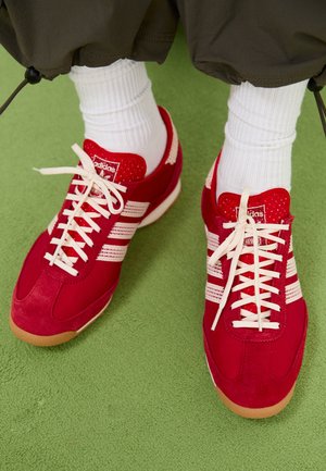 Baskets de sport en suède rouge et mesh avec des rayures et des lacets blancs. Semelle en caoutchouc. Portées avec des chaussettes blanches à côtes sur une surface texturée verte.