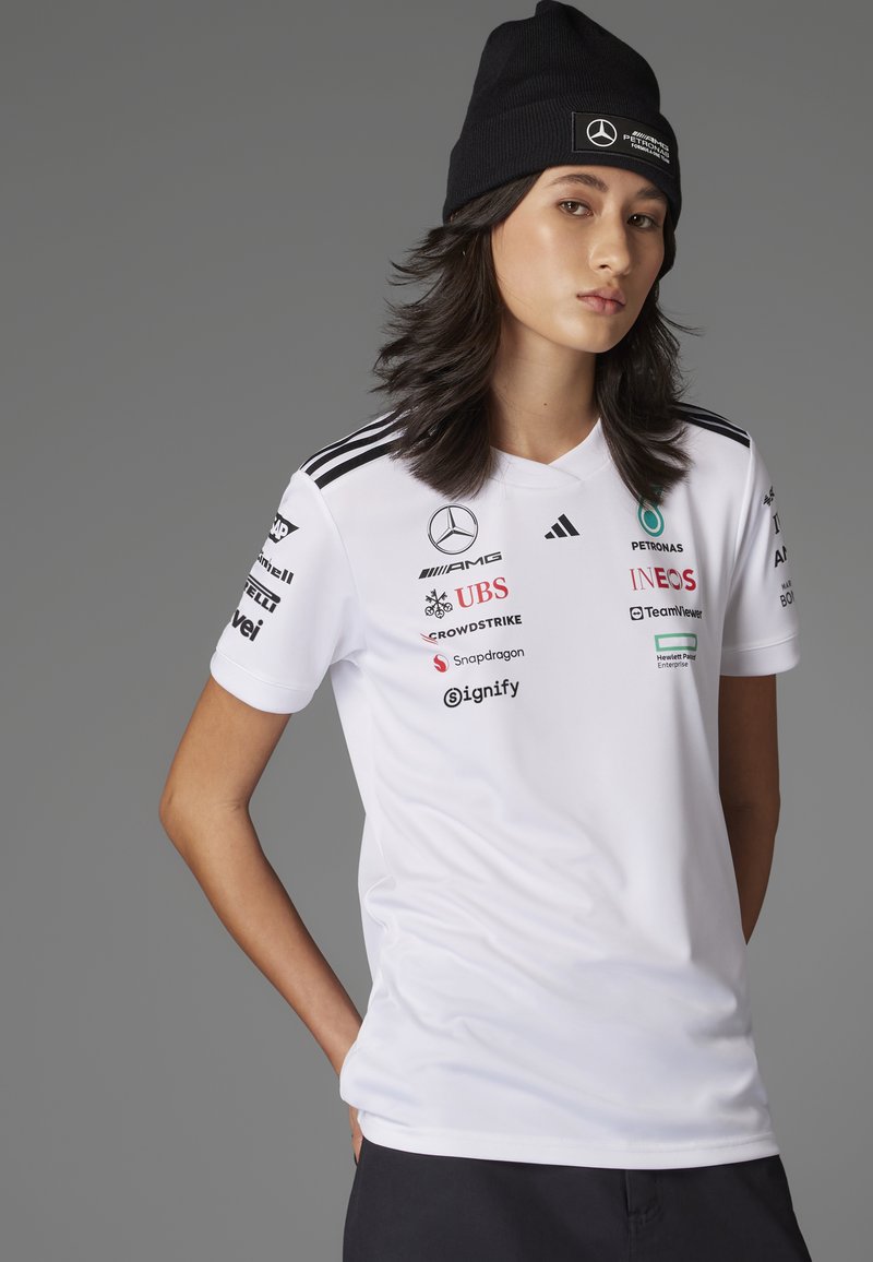 Camiseta deportiva blanca con cuello en V, mangas cortas, rayas negras en los hombros y múltiples logotipos de marcas, incluyendo Mercedes y Petronas.