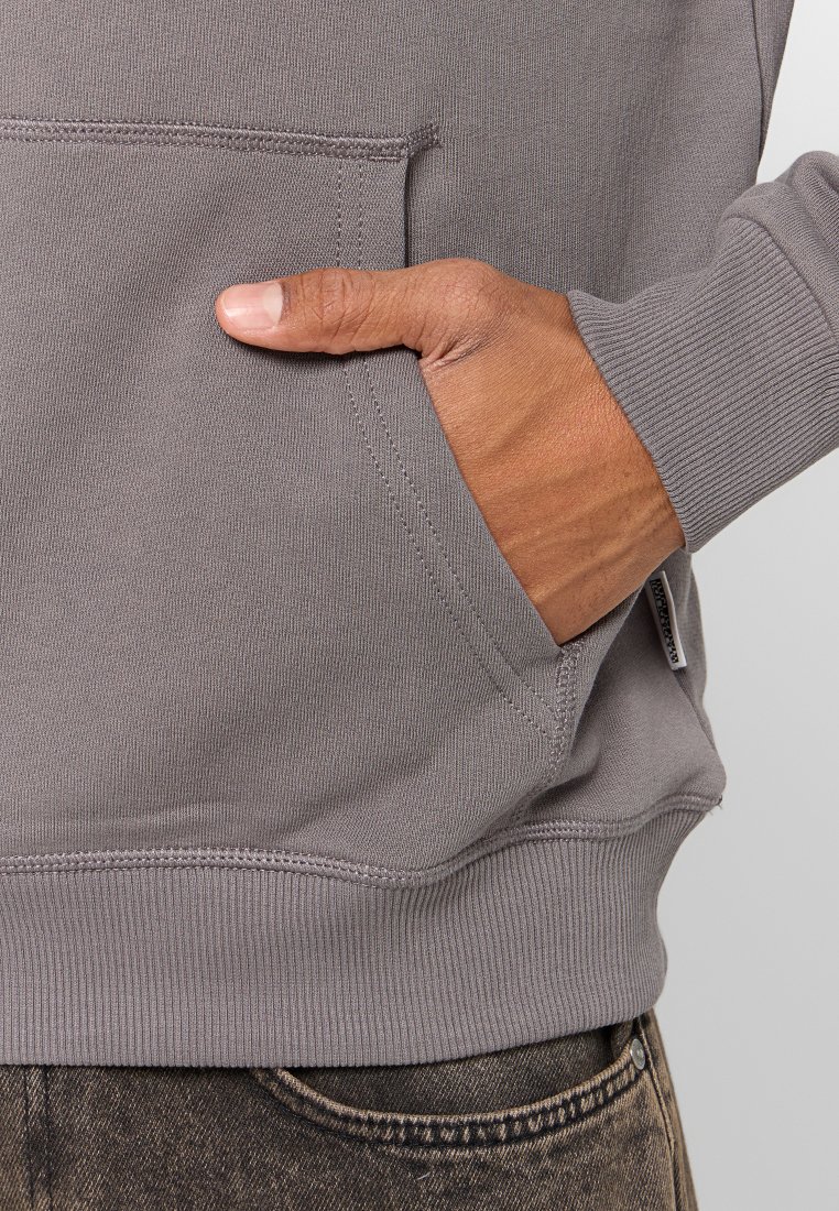 Felpa grigia con polsini e orlo a coste, dotata di una tasca frontale. Realizzata in materiale morbido, con una mano appoggiata nella tasca.