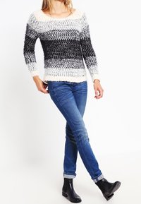 Chunky Strickpullover mit grauen, schwarzen und cremefarbenen horizontalen Streifen; kombiniert mit enganliegenden blauen Jeans und schwarzen Ankle Boots.