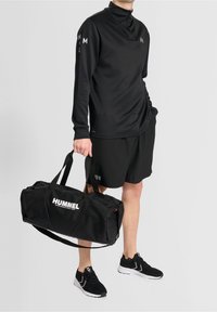 Czarna koszulka sportowa z długim rękawem i wysokim kołnierzem, czarne szorty, czarne buty sportowe oraz czarna torba podróżna z białym logo "HUMMEL".