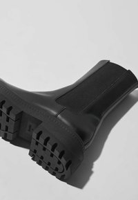 Botte Chelsea en cuir noir avec semelle épaisse à crampons et panneau latéral élastique, montrée couchée sur le côté sur un fond uni.