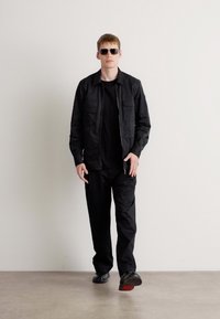 Black lightweight jacket with zip closure, matching black top and pants, paired with black sunglasses and black shoes. Smooth texture throughout.