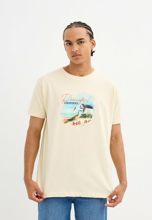 Camiseta de algodón color crema con un estampado gráfico de una casa de playa y el texto "California Pacífica" en tonos naranjas y azules.