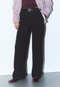 Svarta byxor med vida ben och en slät textur, hög midja, matchat med en randig röd och vit skjorta samt mörkbruna skor.