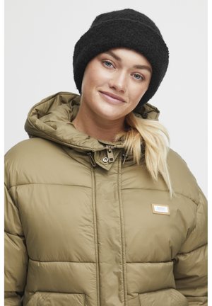 Pufjakke i olivengrøn med hætte, der har et syet mønster og frontlommer. Bæres med en sort strikket beanie.