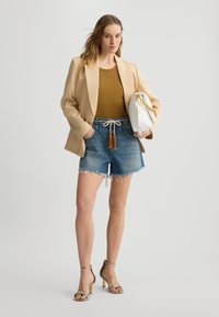 Blazer beige sopra un top marrone, abbinato a pantaloni shorts in denim sfilacciati e tacchi in pelle di serpente. Borsa bianca con dettaglio in catena dorata.