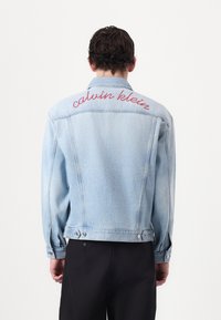 Vaaleansininen farkkutakki, jossa on selkeä punainen brodeerattu "calvin klein" -teksti takana, tavallinen kaulus ja nappikiinnityksellä varustetut hihansuut.