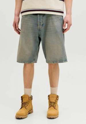 Uomo con pantaloncini di jeans sbiaditi, calzini beige e stivali da lavoro beige con lacci, in piedi contro uno sfondo chiaro e semplice.
