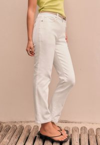 White denim pants with a straight leg cut, featuring front pockets and a belt loop. Worn with a green top and black flip-flops.