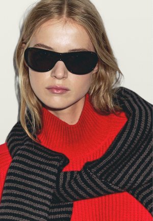 Occhiali da sole neri, dolcevita rosso melange spesso a maglia grossa e sciarpa a righe nere e grigie drappeggiata sulle spalle. Texture liscia, design moderno.