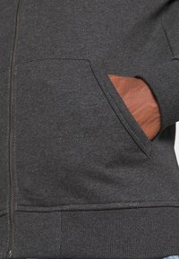 Mano nella tasca anteriore di una felpa grigia scura con zip, dotata di polsini e vita a coste.