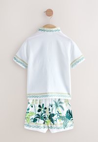 Balti trumpomis rankovėmis marškiniai su žaliu raštuotu apvadu, derinami su šortais, kuriuose yra žalias botanikos raštas ir dekoratyvus juosmens diržo detalizavimas.
