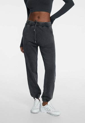 Pantaloni della tuta grigio scuro con vita e polsini elasticizzati, caratterizzati da un dettaglio con coulisse. Abbinati a sneakers bianche su uno sfondo neutro.