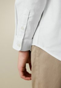 Weißer Hemdärmelschlitz mit Knopf, Seitenansicht, kombiniert mit beiger Hose. Glatte Stofftextur, abgerundeter Ärmelabschluss mit dekorativer Nahtverzierung.