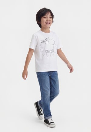 Garçon souriant marchant, portant un t-shirt blanc avec un dessin de chien cartoon, un jean bleu et des baskets noires sur un fond blanc uni.