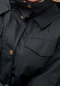 Veste noire avec poche à rabat et boutons pressions dorés, montrant le buste supérieur ainsi qu'une partie du col et du cou.