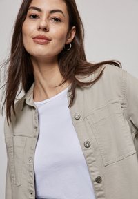 Ung kvinna med långt brunt hår, som bär stora örhängen, en beige knappjacka över en vit t-shirt, framför en ljus neutral bakgrund.