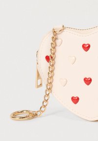 Sac en cuir rose pastel en forme de cœur avec des accents de cœurs rouges et blancs, bandoulière en chaîne dorée et attache porte-clés. Fermeture éclair.