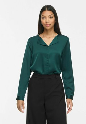 Femme aux longs cheveux foncés portant un chemisier en satin vert foncé et un pantalon noir taille haute, debout sur un fond blanc uni.