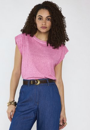 Donna con capelli ricci che indossa un top rosa senza maniche, jeans blu a vita alta, cintura marrone e braccialetti e orecchini dorati.