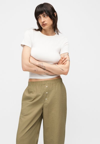 Γυναίκα με κοντά σκούρα μαλλιά και τατουάζ, σταυρωμένα χέρια, φοράει λευκό εφαρμοστό κοντομάνικο crop top και χαλαρό ελαι πράσινο παντελόνι με κουμπιά.