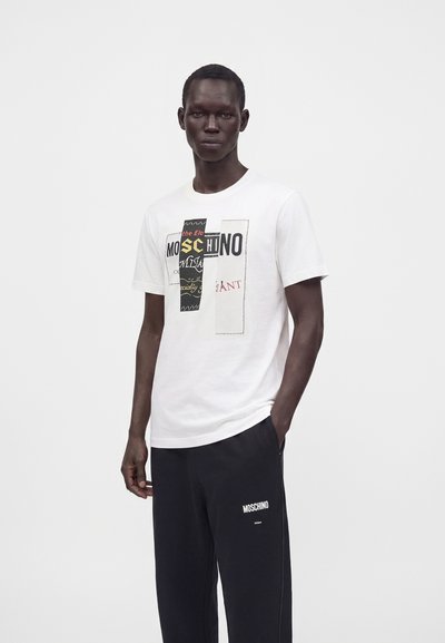 Άνδρας που φοράει λευκό t-shirt Moschino με στάμπα και μαύρη φόρμα Moschino, στέκεται με το ένα χέρι στην τσέπη πάνω σε μονόχρωμο φόντο.