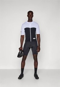 Άνδρας ποδηλάτης με μαύρη και λευκή εφαρμοστή φανέλα και σορτσάκι ποδηλασίας, κρατώντας μαύρο κράνος, στέκεται μπροστά σε απλό φωτεινό φόντο.