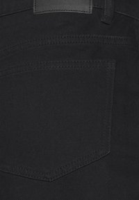 Os jeans de denim preto apresentam um bolso traseiro com uma aba pontiaguda, detalhes cosidos e um tecido texturizado. Uma pequena etiqueta de couro é visível acima da linha da cintura.